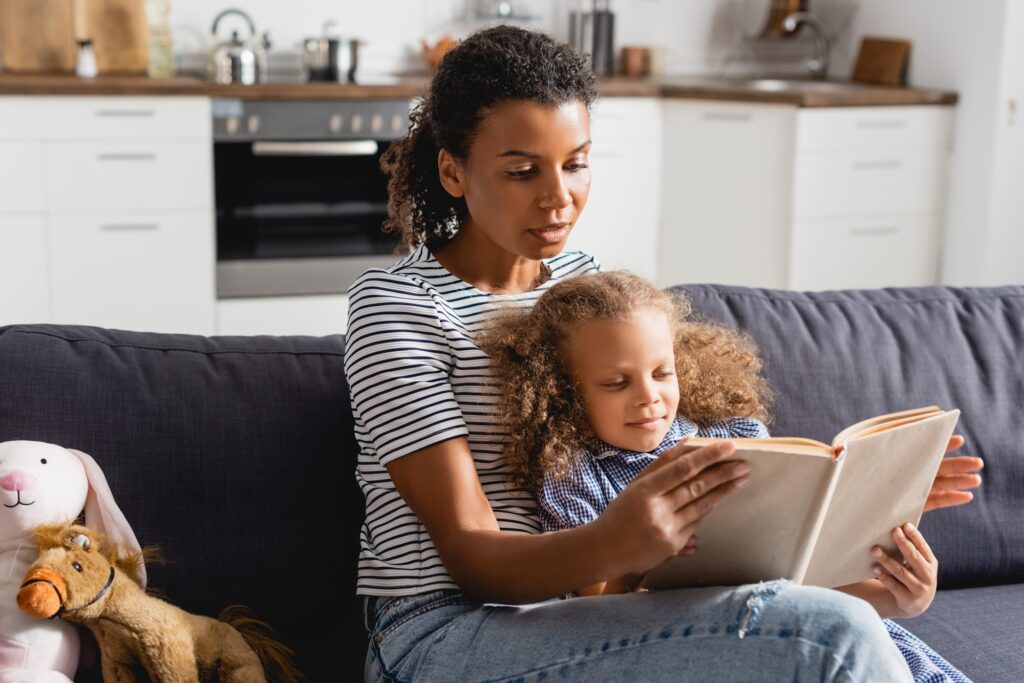 african american girl and young babysitter in striped t-shirt reading book in kitchen